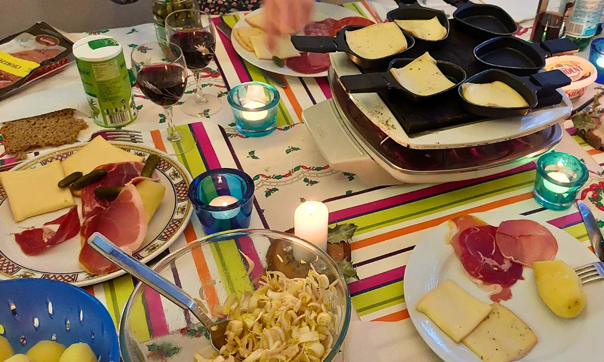 Raclette meal on a colorful table with a raclette machine, melted cheese trays, potatoes, charcuterie, pickles, salad, and candles.